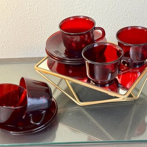 Könnte beinhalten: Ein Set aus vier Tassen und Untertassen aus durchscheinendem rubinrotem Glas. Zwei Tassen stehen auf einem geometrischen, goldfarbenen Metallständer mit Spiegelboden, während die anderen zwei Tassen und Untertassen separat gestapelt sind.