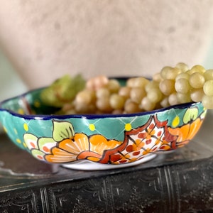 May include: A green ceramic bowl with a scalloped rim and colorful floral designs. The bowl is filled with green grapes and is sitting on a dark brown book.