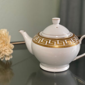 May include: White teapot with a gold Greek key pattern around the middle. The teapot has a lid and a handle.