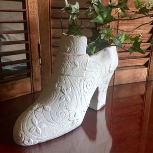 May include: White ceramic planter shaped like a high-heeled shoe with a floral design. The planter is filled with green ivy.