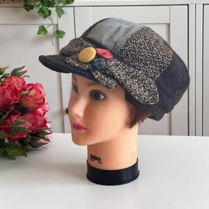 May include: A multi-textured newsboy cap featuring a gray crown, black and tan tweed side panels, and a dark brim. The front is decorated with a fabric bow detail, a round yellow button, and a smaller round red button centered above the brim.