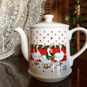 May include: White ceramic teapot with a red, green, and white design of geese. The teapot has a snowflake pattern and a red ribbon.
