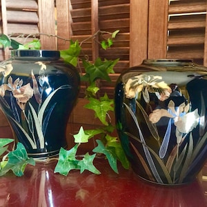 May include: Two black ceramic vases with gold and silver iris flower designs. The vases are on a wooden surface with green ivy.