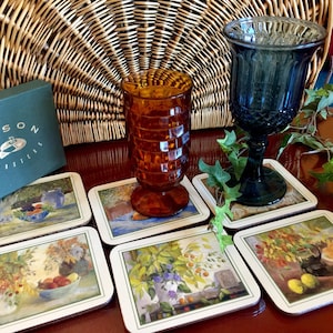 Può includere: Sei sottobicchieri quadrati con illustrazioni colorate di fiori e frutta. I sottobicchieri sono in sughero e hanno un bordo verde. Due bicchieri sono sopra i sottobicchieri. Un bicchiere è ambrato e l'altro è blu.