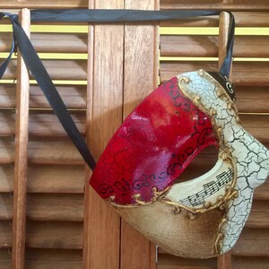 May include: A red and gold masquerade mask with a cracked white design and musical notes. The mask is hanging from a black ribbon.