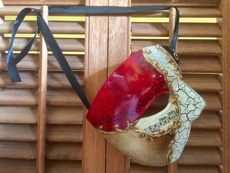 May include: A red and gold masquerade mask with a cracked white design and musical notes. The mask is hanging from a black ribbon.