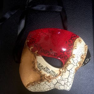 May include: A red, gold and white masquerade mask with a cracked paint effect. The mask has a black ribbon attached and features musical notes on the lower half.