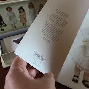 Puede incluir: Una colecci&oacute;n de mu&ntilde;ecas vintage con una mu&ntilde;eca de porcelana con un vestido blanco y zapatos negros. La mu&ntilde;eca est&aacute; de pie sobre un fondo blanco. El texto en la tarjeta dice "Simon y Halbig fue una f&aacute;brica de porcelana en Alemania fundada en 1839. Comenzaron a producir mu&ntilde;ecas en 1800. Simon y Halbig produjeron una gran cantidad de moldes. Tambi&eacute;n produjeron cabezas de mu&ntilde;ecas para otros fabricantes y el mercado franc&eacute;s."
