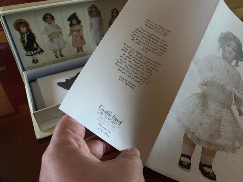 Puede incluir: Una colecci&oacute;n de mu&ntilde;ecas vintage con una mu&ntilde;eca de porcelana con un vestido blanco y zapatos negros. La mu&ntilde;eca est&aacute; de pie sobre un fondo blanco. El texto en la tarjeta dice "Simon y Halbig fue una f&aacute;brica de porcelana en Alemania fundada en 1839. Comenzaron a producir mu&ntilde;ecas en 1800. Simon y Halbig produjeron una gran cantidad de moldes. Tambi&eacute;n produjeron cabezas de mu&ntilde;ecas para otros fabricantes y el mercado franc&eacute;s."