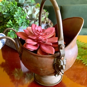 May include: A copper coal scuttle with a lion head handle and a porcelain knob. The scuttle is filled with a pink succulent plant.