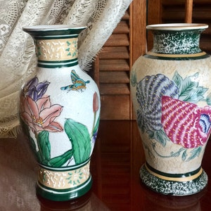 May include: Two ceramic vases with floral and butterfly designs. The taller vase is white with green, pink, and blue flowers and a blue butterfly. The shorter vase is beige with green, pink, and blue flowers.