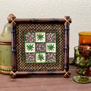 Puede incluir: Un posavasos de bambú marrón con un diseño de azulejos verdes y blancos. El diseño de los azulejos presenta un patrón repetitivo de hojas verdes y guijarros blancos y marrones.