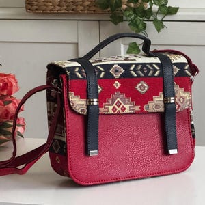 May include: A red crossbody bag with a patterned flap and a textured lower section. The flap features a geometric design in red, black, and gold. The bag has a black handle and straps.
