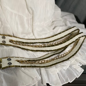May include: Two white leather belts with gold chain accents. The belts are layered on top of each other and have a rectangular pattern.