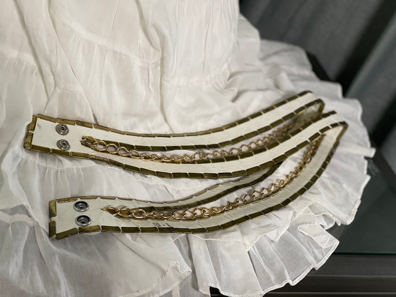 May include: Two white leather belts with gold chain accents. The belts are layered on top of each other and have a rectangular pattern.
