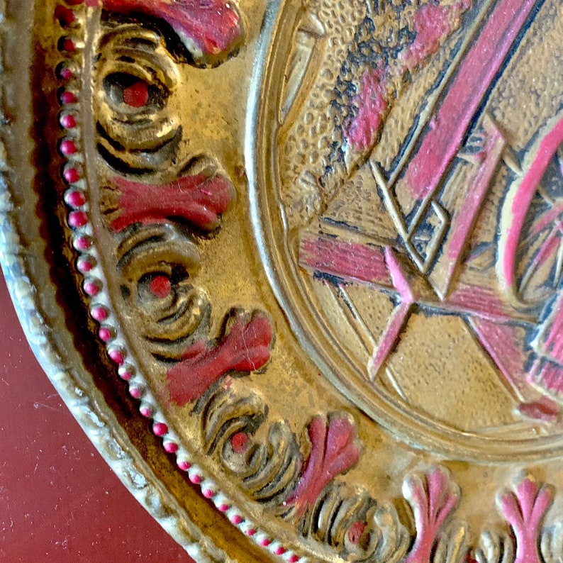 K&ouml;nnte beinhalten: Ein runder, goldfarbener Teller mit einem erhabenen Design. Der Teller hat einen roten und wei&szlig;en Rand und ein erhabenes Design in der Mitte. Das mittlere Design ist ein stilisiertes Bild eines Geb&auml;udes mit einem Rad.