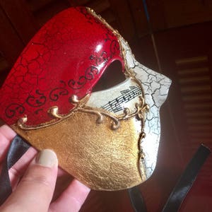 May include: A red, gold and white masquerade mask with a cracked paint design. The mask has a gold trim and musical notes on the white side.