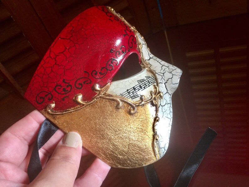 May include: A red, gold and white masquerade mask with a cracked paint design. The mask has a gold trim and musical notes on the white side.