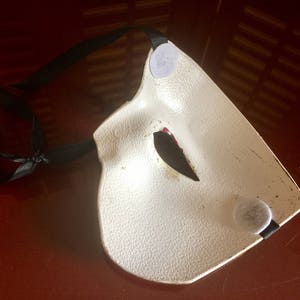 May include: A white masquerade mask with black ribbon ties and a red and black eye design. The mask has two grey felt circles on the back.