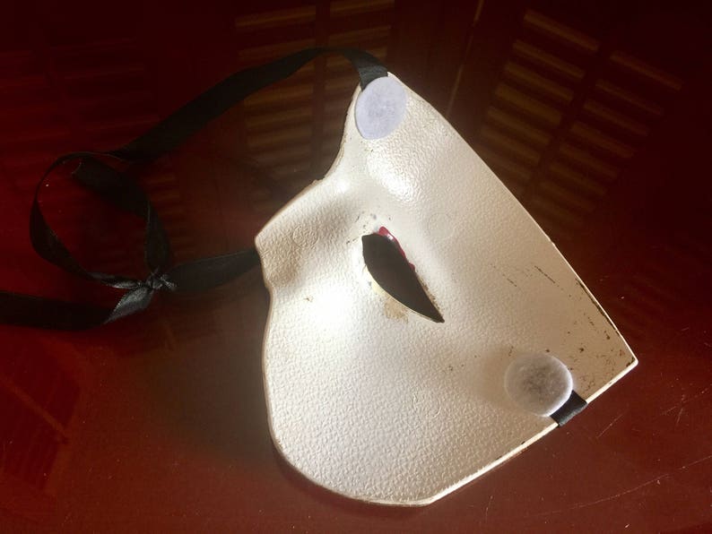 May include: A white masquerade mask with black ribbon ties and a red and black eye design. The mask has two grey felt circles on the back.