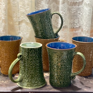 May include: Five ceramic mugs with blue rims and intricate green and brown patterns. The mugs are stacked and arranged on a wooden surface.