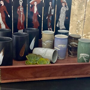 May include: A wooden tray with eight ceramic teacups. The cups are in various colors and designs, including black, white, green, blue, and brown. Some of the cups have a brushstroke design, while others have a more intricate pattern. There is a small green succulent plant in the center of the tray.