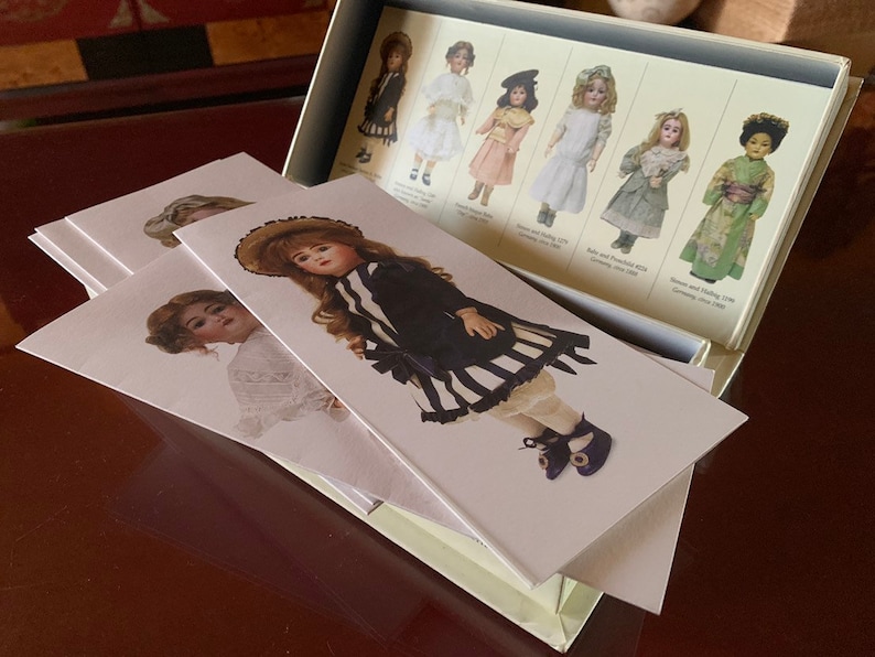 Puede incluir: Un conjunto de cinco tarjetas blancas con im&aacute;genes de mu&ntilde;ecas antiguas. Cada tarjeta presenta una mu&ntilde;eca diferente vestida con una variedad de estilos de ropa. Las mu&ntilde;ecas est&aacute;n todas vestidas con ropa vintage y tienen una variedad de peinados. Las tarjetas est&aacute;n en blanco en la parte posterior y son perfectas para escribir notas o mensajes.