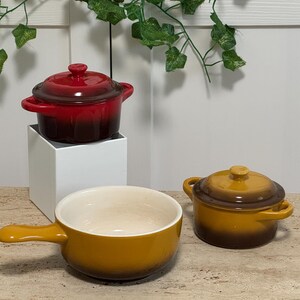 May include: A set of three ceramic cookware pieces including a deep red lidded pot, a mustard yellow lidded pot, and a mustard yellow handled bowl. Each piece features a smooth, glossy finish with a darker gradient towards the base.