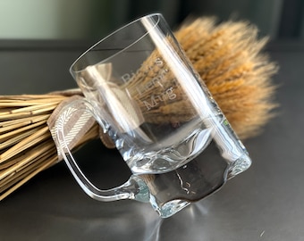 May include: A clear glass beer mug with a handle. The mug is engraved with the words "Dad's Beer Mug".