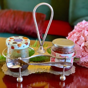 May include: A silver serving stand with two glass jars of jam. The jar on the left has a green and white lid with the text "Menz Gasser". The jar on the right has a silver lid and a red label with the text "Strawberry Fruit Spread".
