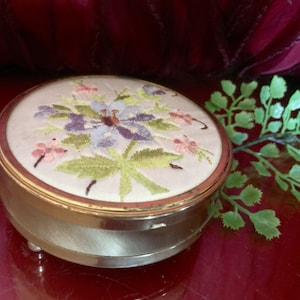 Op de afbeelding: Een vintage, ronde, goudkleurige sieradendoos met een geborduurd bloemenmotief op de deksel. De borduurwerk toont roze, paarse en groene bloemen op een witte achtergrond.