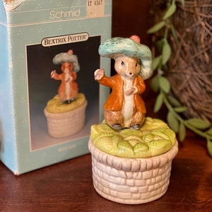Puede incluir: Una caja de música de Beatrix Potter con una figurita de cerámica de Peter Rabbit con un sombrero azul y una chaqueta marrón. La caja está decorada con hojas verdes y una base de mimbre marrón.