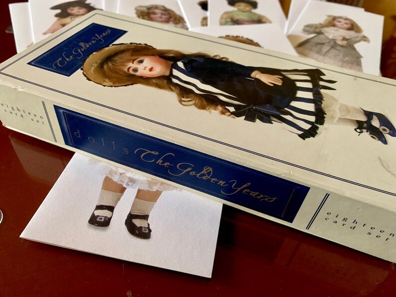 Puede incluir: Una caja de 18 tarjetas coleccionables de mu&ntilde;ecas titulada "Dolls The Golden Years". La caja presenta una mu&ntilde;eca vintage con un vestido a rayas azules y blancas y un sombrero blanco.