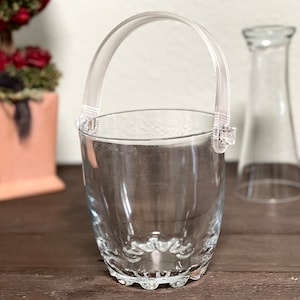 Puede incluir: Un cubo de hielo de plástico transparente con asa. El cubo está vacío y tiene un fondo texturizado.