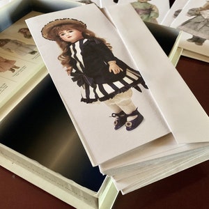 Puede incluir: Una pila de tarjetas en blanco con una ilustraci&oacute;n de mu&ntilde;eca en la tarjeta superior. La mu&ntilde;eca lleva un vestido a rayas blancas y negras con una chaqueta negra y un sombrero marr&oacute;n. La mu&ntilde;eca tiene el pelo rubio y los ojos azules.