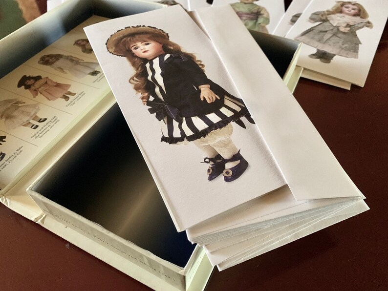 Puede incluir: Una pila de tarjetas en blanco con una ilustraci&oacute;n de mu&ntilde;eca en la tarjeta superior. La mu&ntilde;eca lleva un vestido a rayas blancas y negras con una chaqueta negra y un sombrero marr&oacute;n. La mu&ntilde;eca tiene el pelo rubio y los ojos azules.