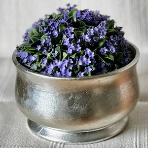 May include: A silver-toned metal bowl filled with vibrant purple flowers and green leaves. The bowl has a wide, rounded shape with a pedestal base. The flowers are clustered together, creating a full, textured appearance. The background is a neutral, textured fabric.