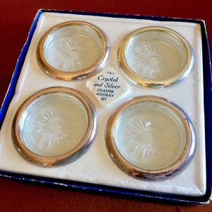Può includere: Set di quattro sottobicchieri e posacenere in vetro con bordo argentato in una scatola bianca con bordo blu. La scatola ha un'etichetta che recita "Italy Crystal and Silver Coaster Ashtray Set".