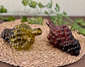 May include: Two glass bottles shaped like bunches of grapes. One bottle is green and the other is purple.