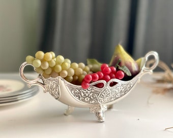 May include: A silver fruit bowl with an ornate design, featuring a green grape cluster and red berries. The bowl has three legs and a handle on each side.