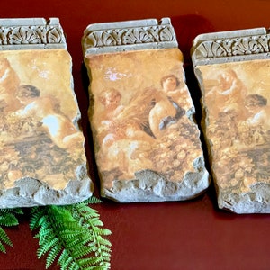 May include: Three decorative stone plaques with a classical painting of cherubs in a garden scene. The plaques have a weathered, textured appearance with a decorative border at the top. The color palette includes beige, brown, and gray.