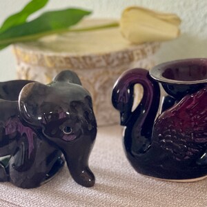 Può includere: Due portacandele in ceramica, uno a forma di elefante e l'altro a forma di cigno. L'elefante è nero e il cigno è viola scuro.