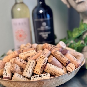 May include: A bamboo bowl filled with used wine corks, with various brand names visible. Two wine bottles are in the background, one white and one black. The corks are a mix of light and dark brown.