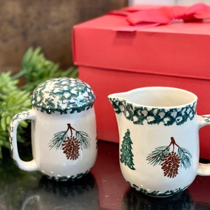Puede incluir: Un juego de crema y azúcar de cerámica blanca con un diseño de pino verde y marrón. El juego está sobre una superficie negra delante de una caja de regalo roja.