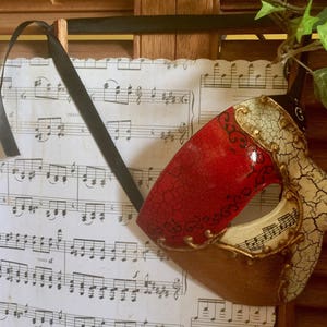 May include: A red and white masquerade mask with gold accents and a cracked, textured finish. The mask is lying on a sheet of music.