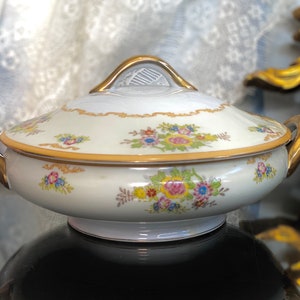 Op de afbeelding: Een witte porseleinen serveerschaal met een gouden rand en een bloemenpatroon. De schaal heeft een deksel met een gouden handvat.