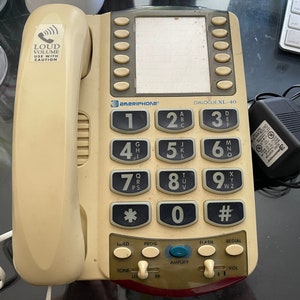 May include: Cream-colored Ameriphone Dialogue XL-40 telephone with large number buttons. Features a speakerphone, volume control, and a power adapter. The phone has a sticker that says "LOUD VOLUME USE WITH CAUTION."