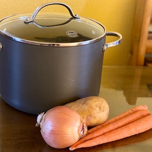 Op de afbeelding: Een grijze pot met een glazen deksel, een bruine ui, een aardappel en drie oranje wortels op een houten tafel.
