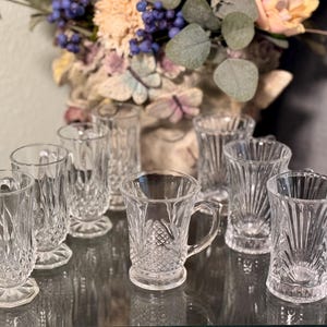 Könnte beinhalten: Ein Set aus acht klaren Glasbechern mit aufwendigen Schnitzereien. Die Becher haben einen Fuß und einen Griff. Die Becher sind auf einem Tisch mit einer reflektierenden Oberfläche angeordnet.