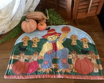 May include: A quilted fabric teapot cosy featuring a scarecrow holding a pumpkin, sitting on a fence with two pumpkins. The cosy is decorated with green, brown, blue, red, and yellow fabric.